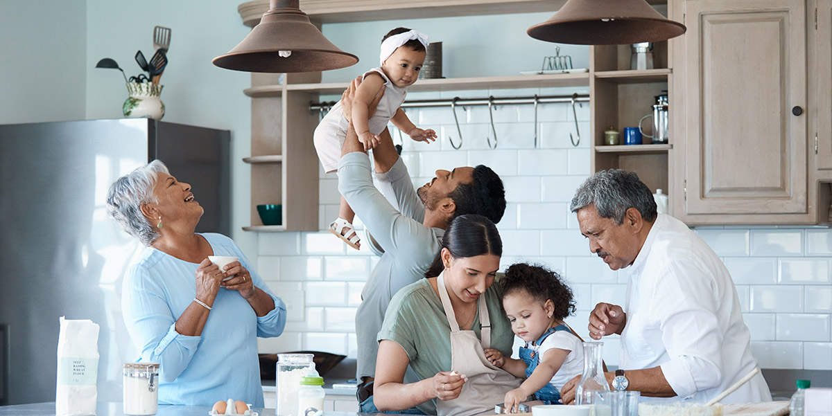 Two parent, 2 kids and 2 grand parents in a kitchen. The father holds the youngest child up in the air.