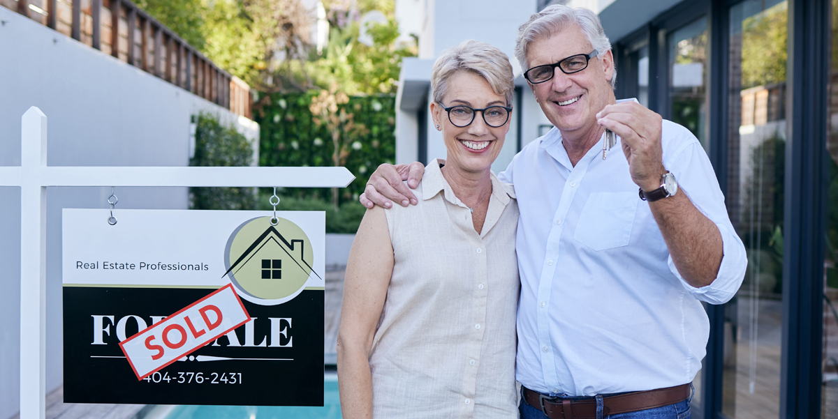 Baby Boomer couple in front of modern home with sales sign after having sold their home.