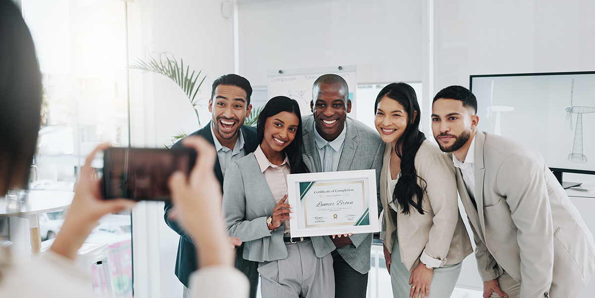 A mixed race and gender team at work holding a certificate and being photographed by a coworker.