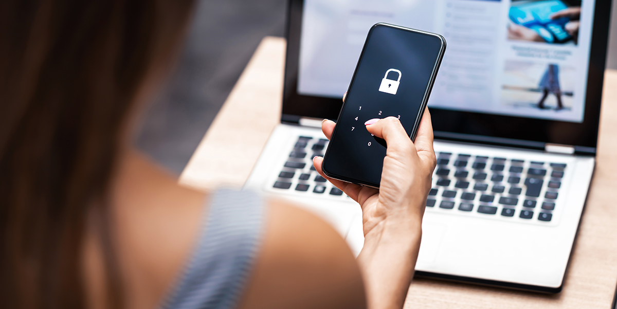 Woman from behind sitting in front of laptop, holding a cellphone with a security lock icon on the screen.