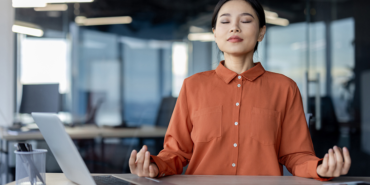 Asian woman sitting at office desk with eyes closed and hands in yoga meditation position.