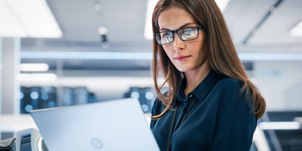 Smart-looking woman with glasses looking at a laptop in an office environment.