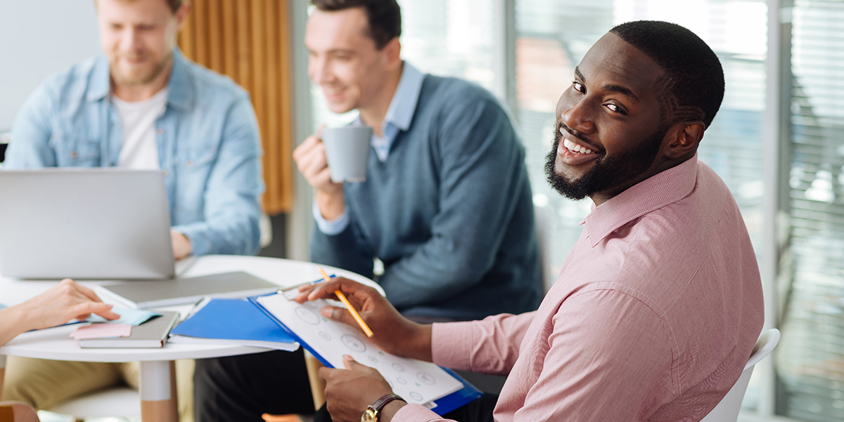 African American male manager smiling into the camera with colleagues working in the background.